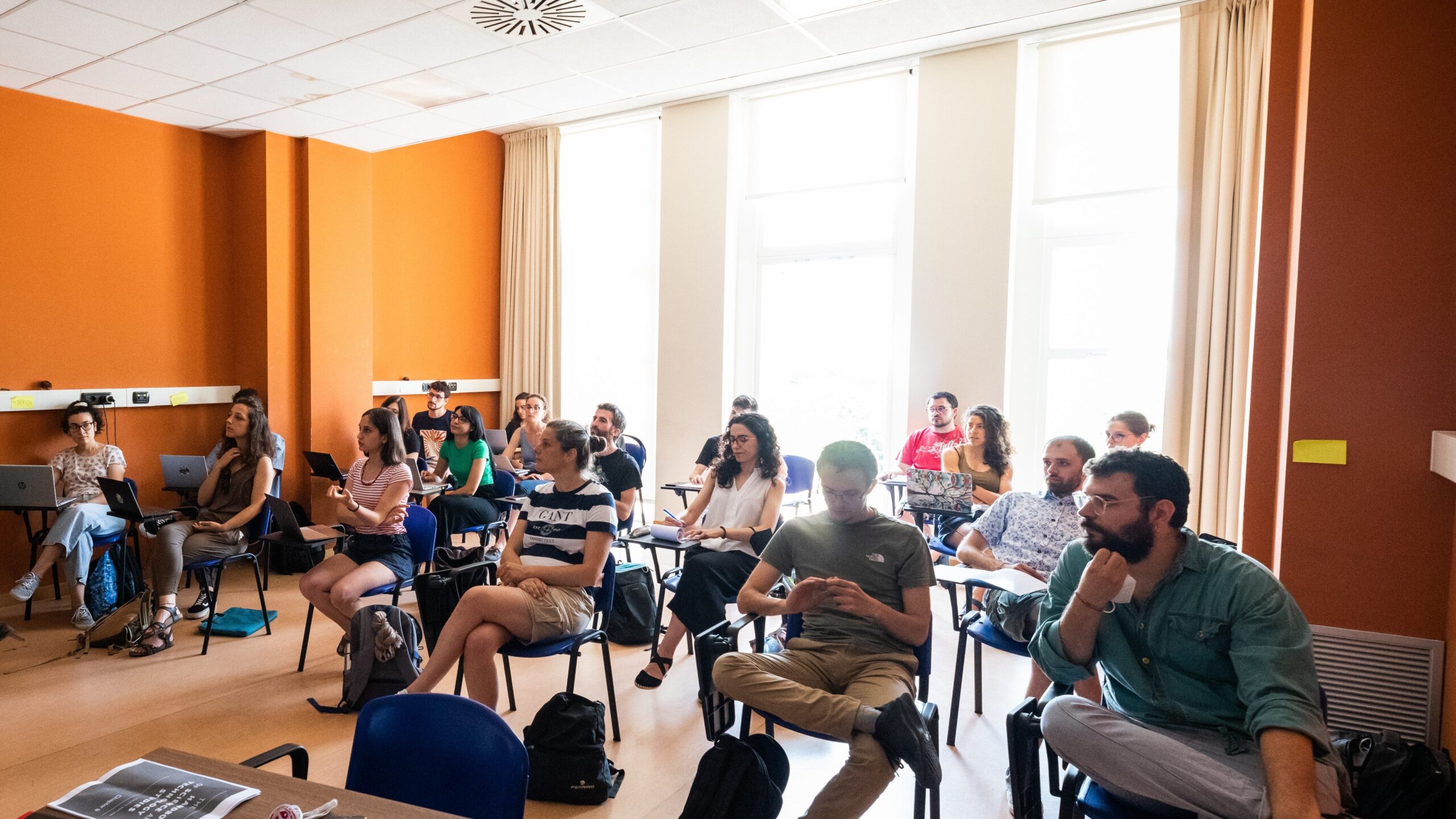 Partecipanti seduti in aula durante una lezione del Master in Comunicazione della Scienza, con pareti arancioni e finestre lu